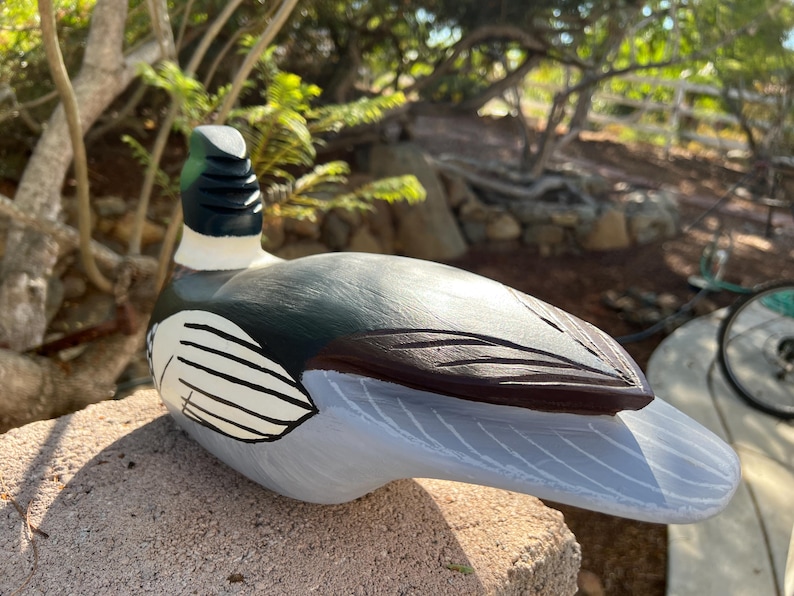 May include: A hand-painted wooden duck figurine. The duck has a green head, black and white neck, and a gray and brown body. The duck is resting on a stone surface.