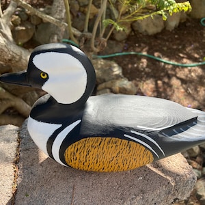 May include: A hand-painted wooden duck figurine. The duck has a white head with black markings, a black beak, and a yellow eye. The body is grey with black and white accents, and a textured yellow and brown section on the side.