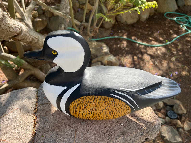 May include: A hand-painted wooden duck figurine. The duck has a white head with black markings, a black beak, and a yellow eye. The body is grey with black and white accents, and a textured yellow and brown section on the side.