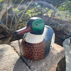 May include: A hand-painted wooden duck figurine. The duck has a green head, a red beak, and a body with a white chest, brown speckled breast, and blue and black back. The duck is sitting on a stone.