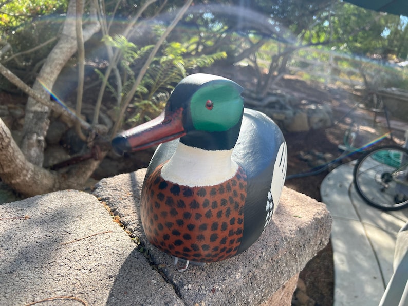 May include: A hand-painted wooden duck figurine. The duck has a green head, a red beak, and a body with a white chest, brown speckled breast, and blue and black back. The duck is sitting on a stone.