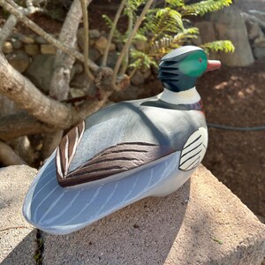 May include: A hand-painted wooden duck figurine with a green head, black and white accents, and a light blue body. The duck has brown wing details and a red beak. The figurine is resting on a stone surface.
