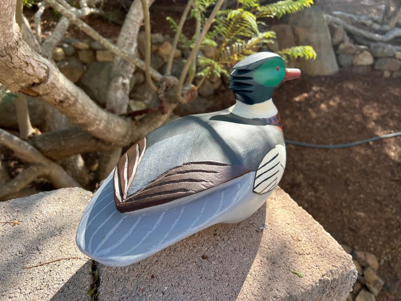 May include: A hand-painted wooden duck figurine with a green head, black and white accents, and a light blue body. The duck has brown wing details and a red beak. The figurine is resting on a stone surface.