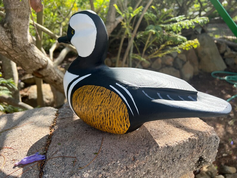 May include: A hand-painted wooden duck decoy. The duck has a black body, white head with a black beak, and a yellow and brown textured chest. The decoy is resting on a stone surface, with greenery in the background.