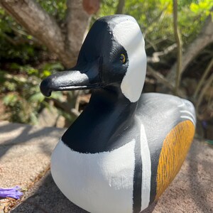 May include: A hand-painted wooden duck ornament. The duck has a black head, white markings, and a body with yellow and grey details. The beak is black. The ornament is displayed on a stone surface.