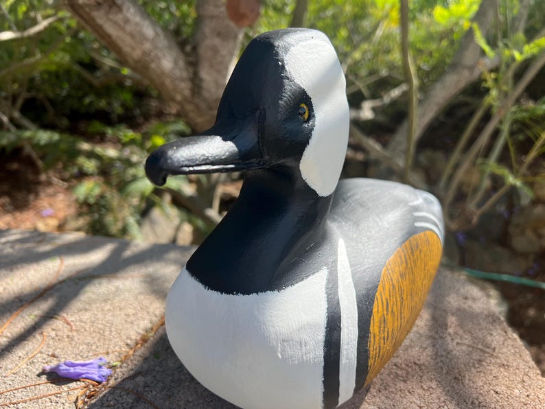 May include: A hand-painted wooden duck ornament. The duck has a black head, white markings, and a body with yellow and grey details. The beak is black. The ornament is displayed on a stone surface.