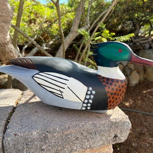 May include: A hand-painted wooden duck sculpture. The duck features a green head with a red eye and beak, a black back, and a white and black wing design. The body has brown and white spots. The duck is resting on a stone surface.