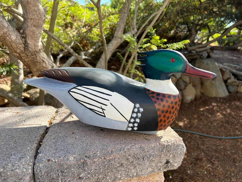 May include: A hand-painted wooden duck sculpture. The duck features a green head with a red eye and beak, a black back, and a white and black wing design. The body has brown and white spots. The duck is resting on a stone surface.