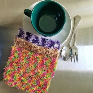 May include: A set of three colorful crocheted dishcloths in a stack. The top dishcloth is a bright orange, yellow, and green color. The middle dishcloth is a brown color. The bottom dishcloth is a purple and white color. The dishcloths are sitting on a silver surface next to a green mug and a white saucer.
