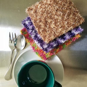 May include: A set of four colorful crocheted dishcloths stacked on top of each other. The dishcloths are made of a soft, fluffy yarn and are perfect for cleaning dishes or wiping down surfaces. The dishcloths are sitting on a white saucer next to a green teacup and two silver forks.