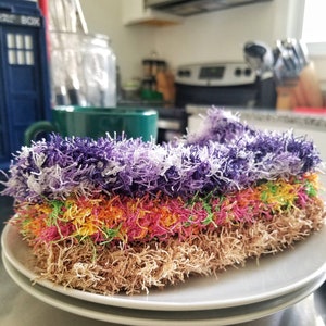 May include: A stack of colorful, fluffy dishcloths in purple, pink, yellow, green, and brown. The dishcloths are arranged in a stack on a white plate.