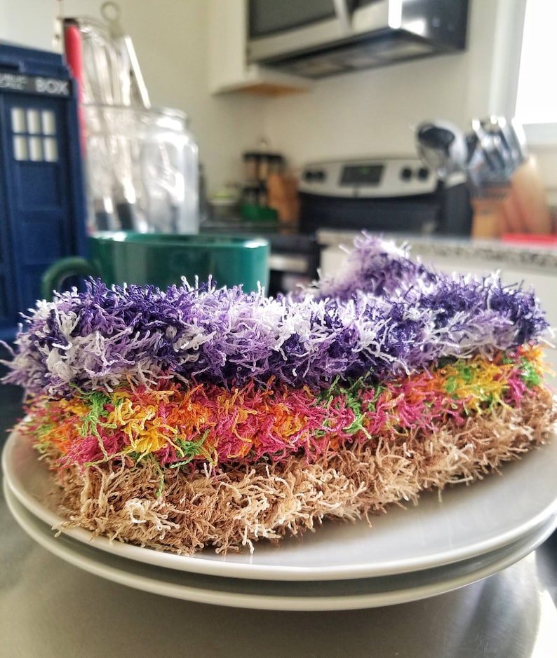May include: A stack of colorful, fluffy dishcloths in purple, pink, yellow, green, and brown. The dishcloths are arranged in a stack on a white plate.