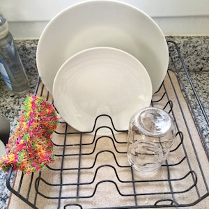 May include: A black metal dish rack with two white ceramic plates and a clear glass jar. A colorful, multi-colored, crocheted dishcloth is in the rack.
