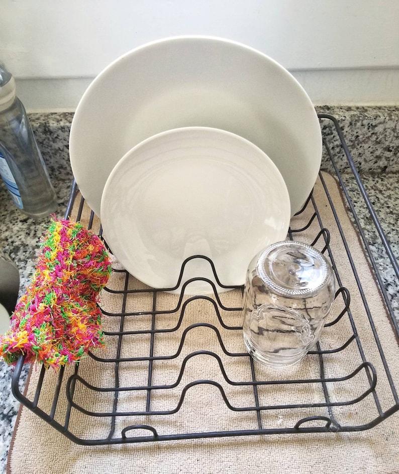 May include: A black metal dish rack with two white ceramic plates and a clear glass jar. A colorful, multi-colored, crocheted dishcloth is in the rack.