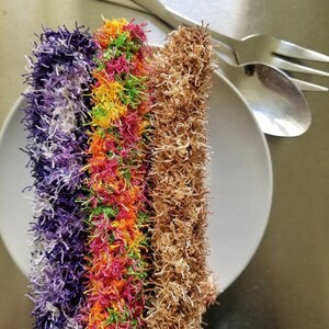 May include: Three colorful, fluffy scrubbers on a white plate. The scrubbers are purple, green and orange with yellow stripes, and brown.