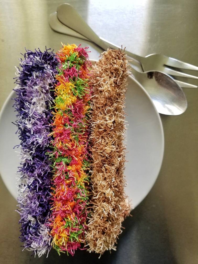 May include: Three colorful, fluffy scrubbers on a white plate. The scrubbers are purple, green and orange with yellow stripes, and brown.