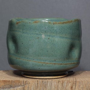 May include: A small, round ceramic bowl with a teal-green glaze and a speckled texture. The bowl features a wavy, uneven rim and several indented thumb-like impressions along its sides, with a visible brown clay base and subtle horizontal lines.