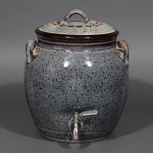 May include: A speckled blue ceramic beverage dispenser with a lid and a spigot. The lid has a handle and decorative cutouts. The dispenser has two handles on the sides. The spigot is made of metal.