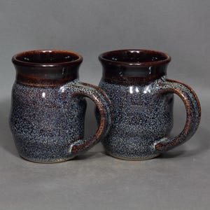 May include: Two ceramic mugs with a speckled blue and brown glaze. The rims and handles are a darker brown, contrasting with the lighter blue speckled body. The mugs have a rounded shape and a sturdy handle, suitable for hot beverages.