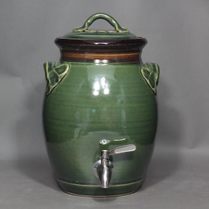 May include: A green ceramic drinks dispenser with a lid and a silver tap. The dispenser has a rounded shape with leaf-shaped handles and a dark brown rim. The lid has a loop handle.