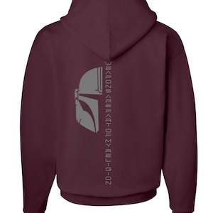 Puede incluir: Sudadera con capucha color burdeos con un gr&aacute;fico de casco mandaloriano gris y el texto "This is the way" en escritura mandaloriana en la parte posterior.