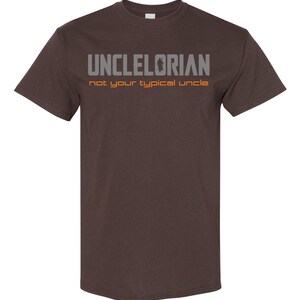 Puede incluir: Una camiseta marr&oacute;n con el texto "UNCLELORIAN not your typical uncle" en letras grises y naranjas.