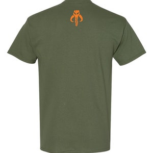 Puede incluir: Una camiseta verde con un s&iacute;mbolo mandaloriano naranja en la espalda.