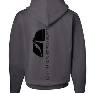 Puede incluir: Una sudadera con capucha gris oscuro con una silueta negra de un casco mandaloriano en la espalda. El texto "This is the way" est&aacute; escrito en escritura mandaloriana en la parte posterior de la sudadera.