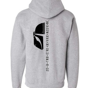Puede incluir: Sudadera con capucha gris con un gr&aacute;fico de casco mandaloriano negro y el texto "Weapons are part of my religion" impreso verticalmente en la espalda.
