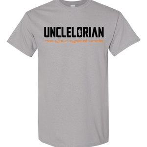 Puede incluir: Una camiseta gris con texto negro que dice "Unclelorian not your typical uncle". El texto est&aacute; en negrita y la palabra "Unclelorian" tiene una peque&ntilde;a silueta de una persona de pie en el medio de la palabra.