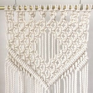 May include: A white macrame wall hanging with a geometric pattern and fringe. The hanging is made with natural cotton rope and is attached to a gold rod.