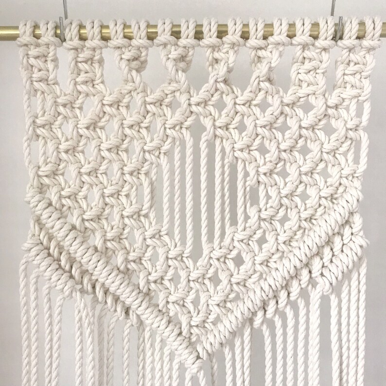 May include: A white macrame wall hanging with a geometric pattern and fringe. The hanging is made with natural cotton rope and is attached to a gold rod.