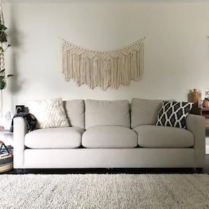 May include: A light grey sofa with three cushions and two throw pillows. One pillow is white and fluffy, the other is black and white with a geometric pattern. The sofa is in front of a white wall with a macrame wall hanging above it.