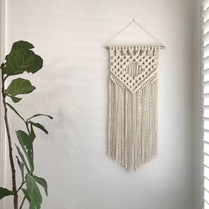 May include: A white macrame wall hanging with a geometric pattern and fringe. The hanging is attached to a wall with a gold rod.