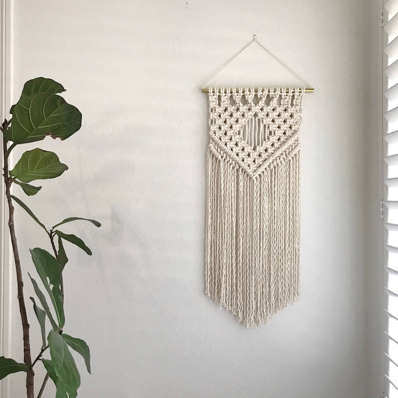 May include: A white macrame wall hanging with a geometric pattern and fringe. The hanging is attached to a wall with a gold rod.