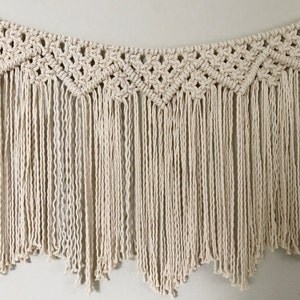 May include: A macrame wall hanging with a geometric pattern and fringe. The hanging is made of natural coloured cotton cord.