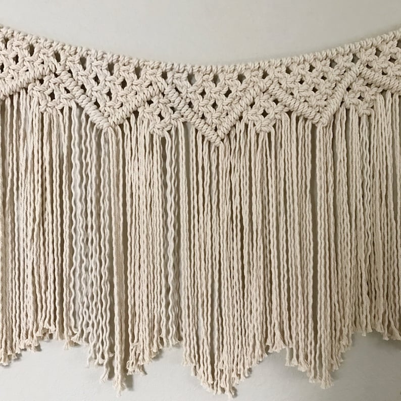 May include: A macrame wall hanging with a geometric pattern and fringe. The hanging is made of natural coloured cotton cord.