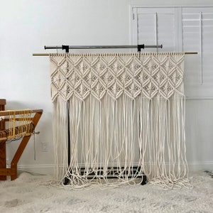 Puede incluir: Un tapiz de macram&eacute; blanco con un patr&oacute;n de diamante, colgado de un soporte de metal negro. El macram&eacute; est&aacute; hecho con cord&oacute;n de algod&oacute;n grueso y tiene una franja en la parte inferior.