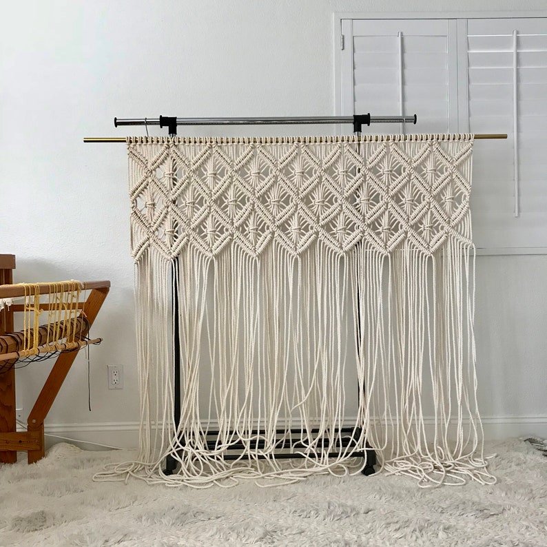 Puede incluir: Un tapiz de macram&eacute; blanco con un patr&oacute;n de diamante, colgado de un soporte de metal negro. El macram&eacute; est&aacute; hecho con cord&oacute;n de algod&oacute;n grueso y tiene una franja en la parte inferior.