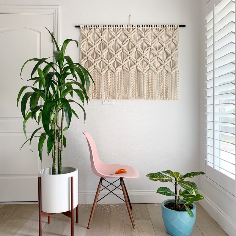May include: A white planter with a green plant sits on a wooden stand next to a pink chair. A macrame wall hanging with a geometric pattern hangs on the wall behind the plant.