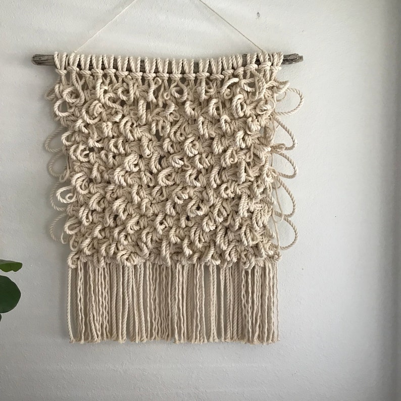 May include: A beige macrame wall hanging with a fringe border. The hanging is made with natural fiber rope and is attached to a driftwood branch.