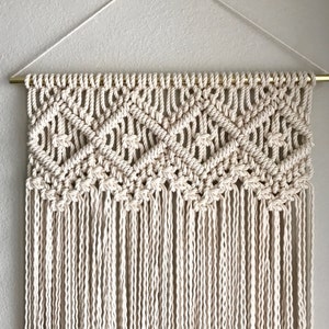 May include: A macrame wall hanging with a geometric pattern and a fringe border. The hanging is made with natural colored cotton cord and is attached to a gold metal rod.