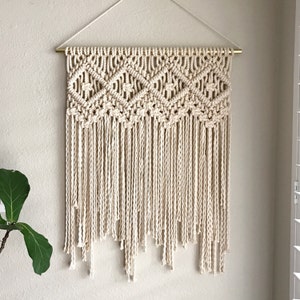 May include: A macrame wall hanging with a geometric pattern and fringe. The hanging is made of natural cotton cord and is attached to a gold metal rod.