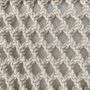 May include: A close-up of a white macrame wall hanging with a diamond pattern. The macrame is made of thick, natural cotton rope.