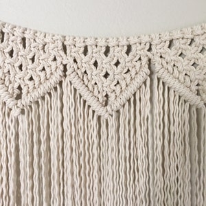 May include: A white macrame wall hanging with a geometric pattern and fringe.