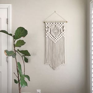 May include: A white macrame wall hanging with a geometric pattern and fringe. The hanging is attached to a gold rod and is hanging on a white wall.