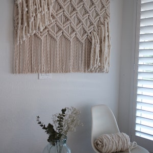 May include: A white macrame wall hanging with a geometric pattern, hanging on a white wall. A white chair with a white yarn roll on it, and a vase with dried flowers on a woven stool.