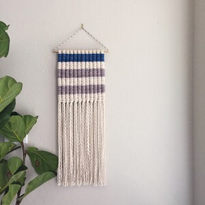 Może przedstawiać: Wiszący gobelin tkany z geometrycznym wzorem białych, szarych i niebieskich pask&oacute;w. Gobelin ma frędzle z białej przędzy.