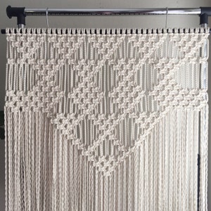 May include: A white macrame wall hanging with a geometric pattern. The hanging is made of cotton cord and is attached to a metal rod.