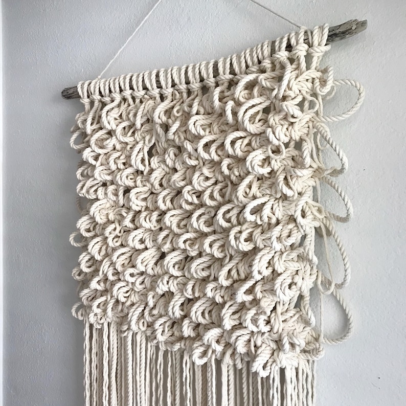 May include: A white macrame wall hanging with a natural wood dowel at the top. The macrame is made with a thick cotton cord and has a fringe at the bottom.